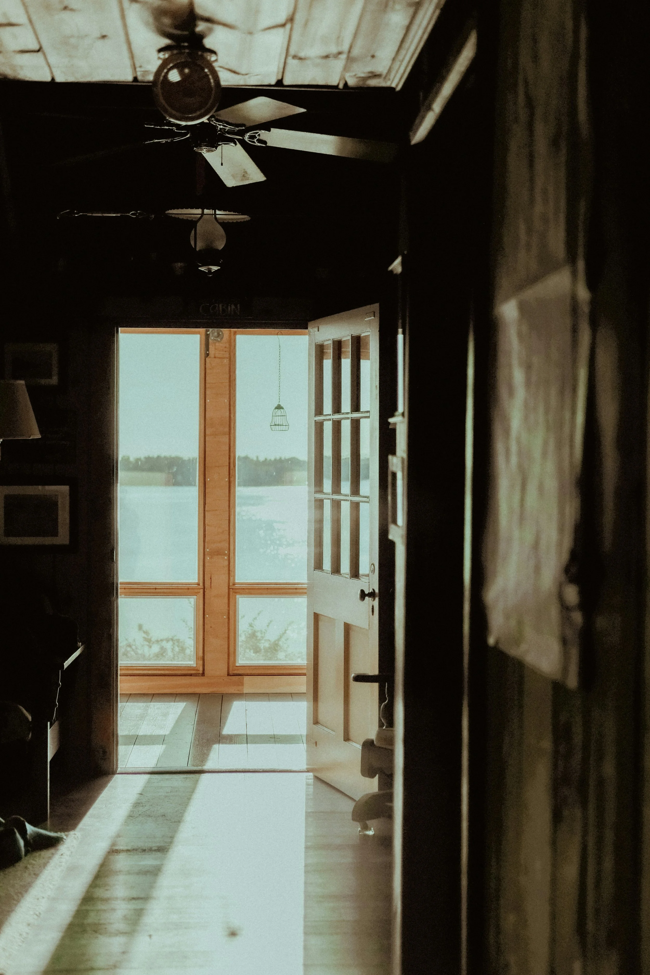 Interior of a house with a partially open front door and sunlight streaming in, revealing a porch with a hanging lamp outside and a scenic view of water and trees in the distance.