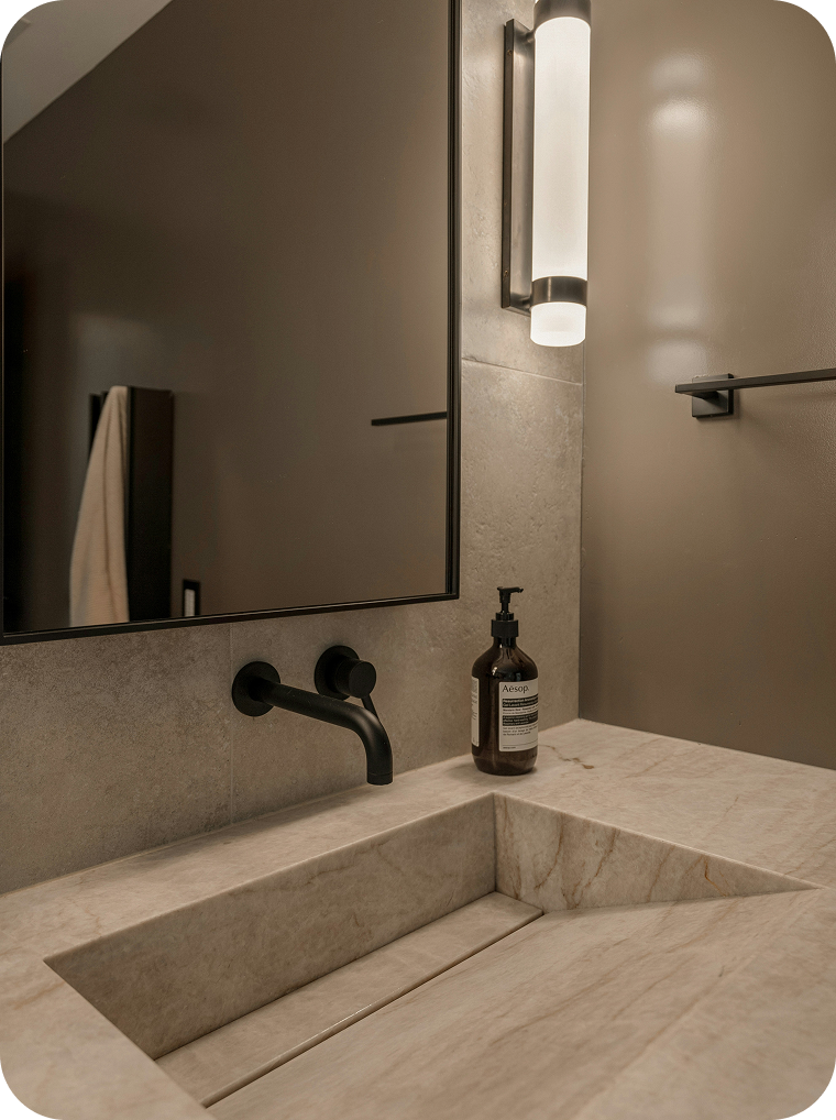 Bathroom vanity with a marble countertop and integrated sink, black wall-mounted faucet, brown soap dispenser, large mirror, and wall-mounted light fixture.