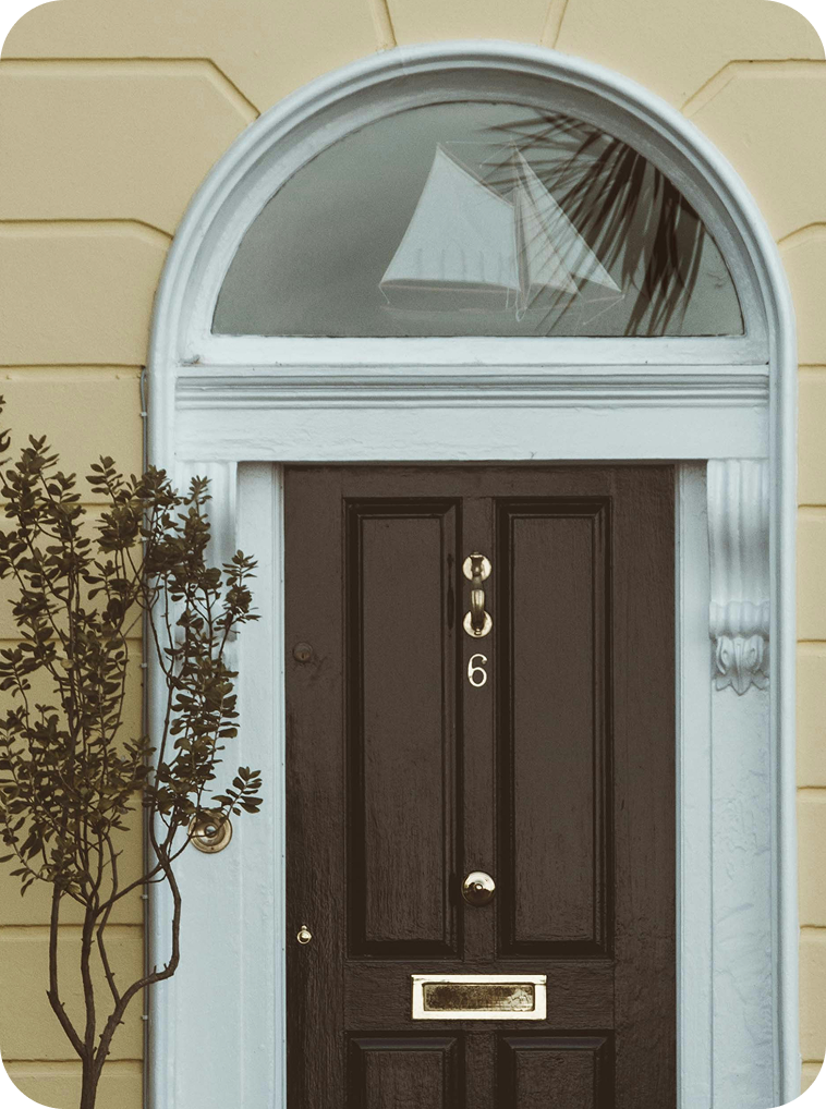 Front door with an arched window and the number 6, flanked by a small shrub, with the reflection of a patio umbrella and palm leaves visible in the window.