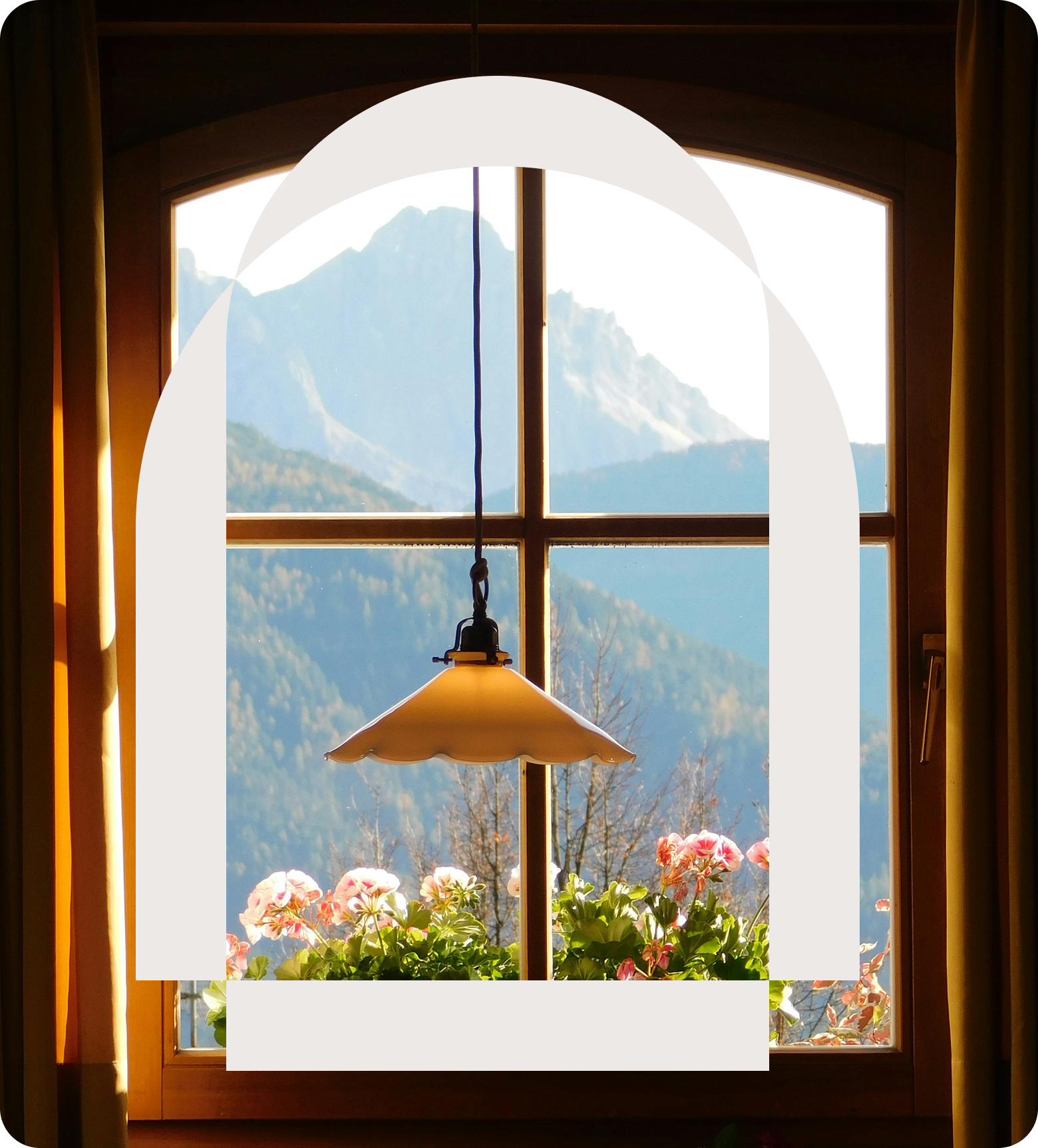 A view through a window showing mountains in the background, a hanging lamp, pink flowers outside, and wooden window frames with curtains.
