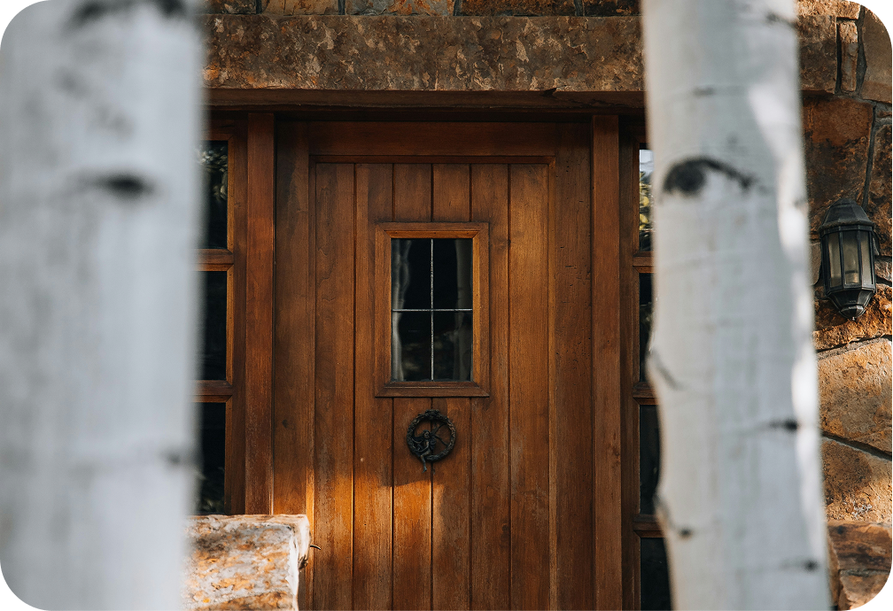 Wooden front door with small window and a decorative wreath, framed by two white painted trees and a stone exterior wall.