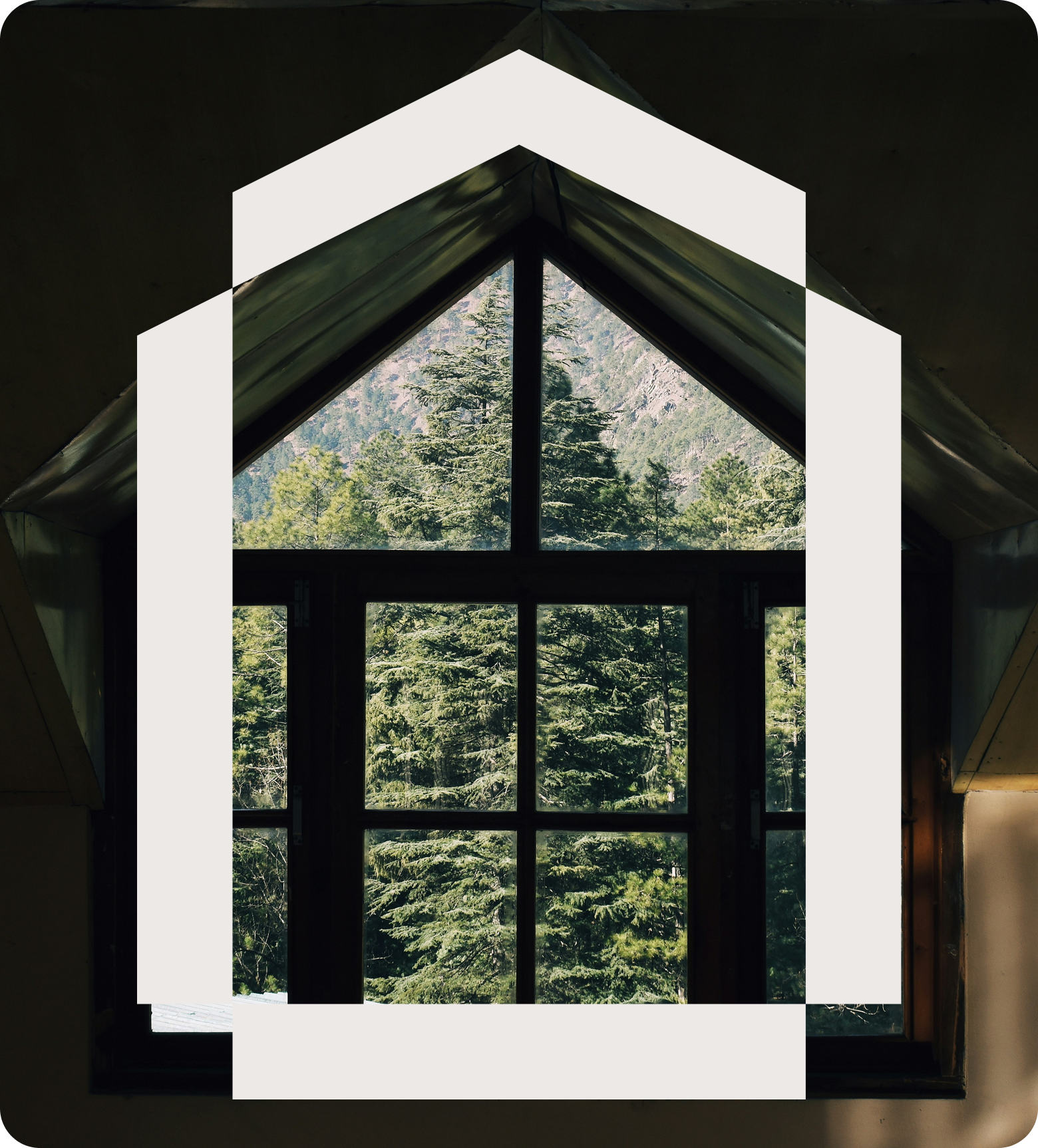 View through a triangular window showing a dense forest of green trees and mountain in the background, with the window frame in the interior of a building.