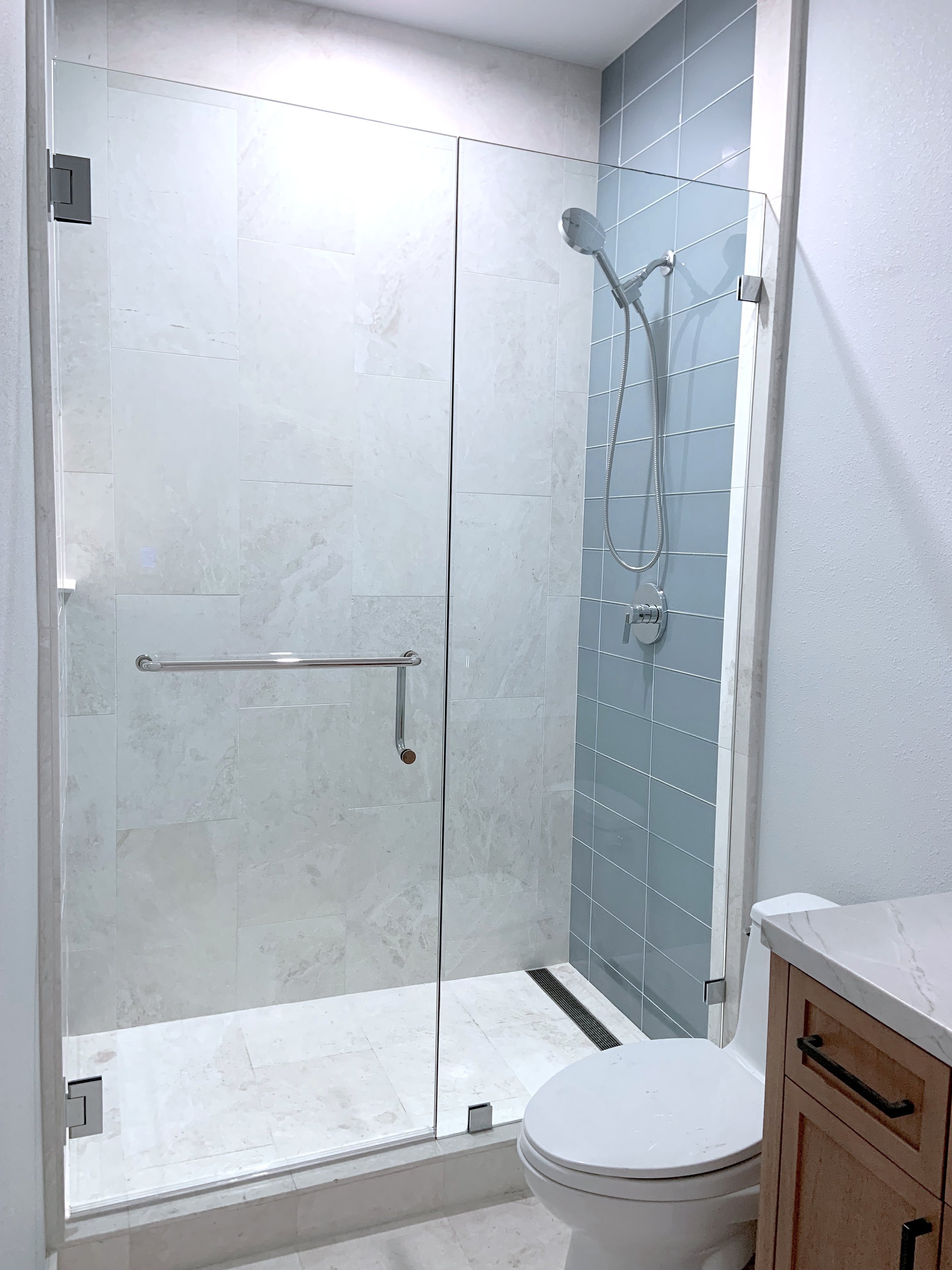 Bathroom with walk-in shower, glass door, gray and white tiles, toilet, and wooden vanity with marble countertop.