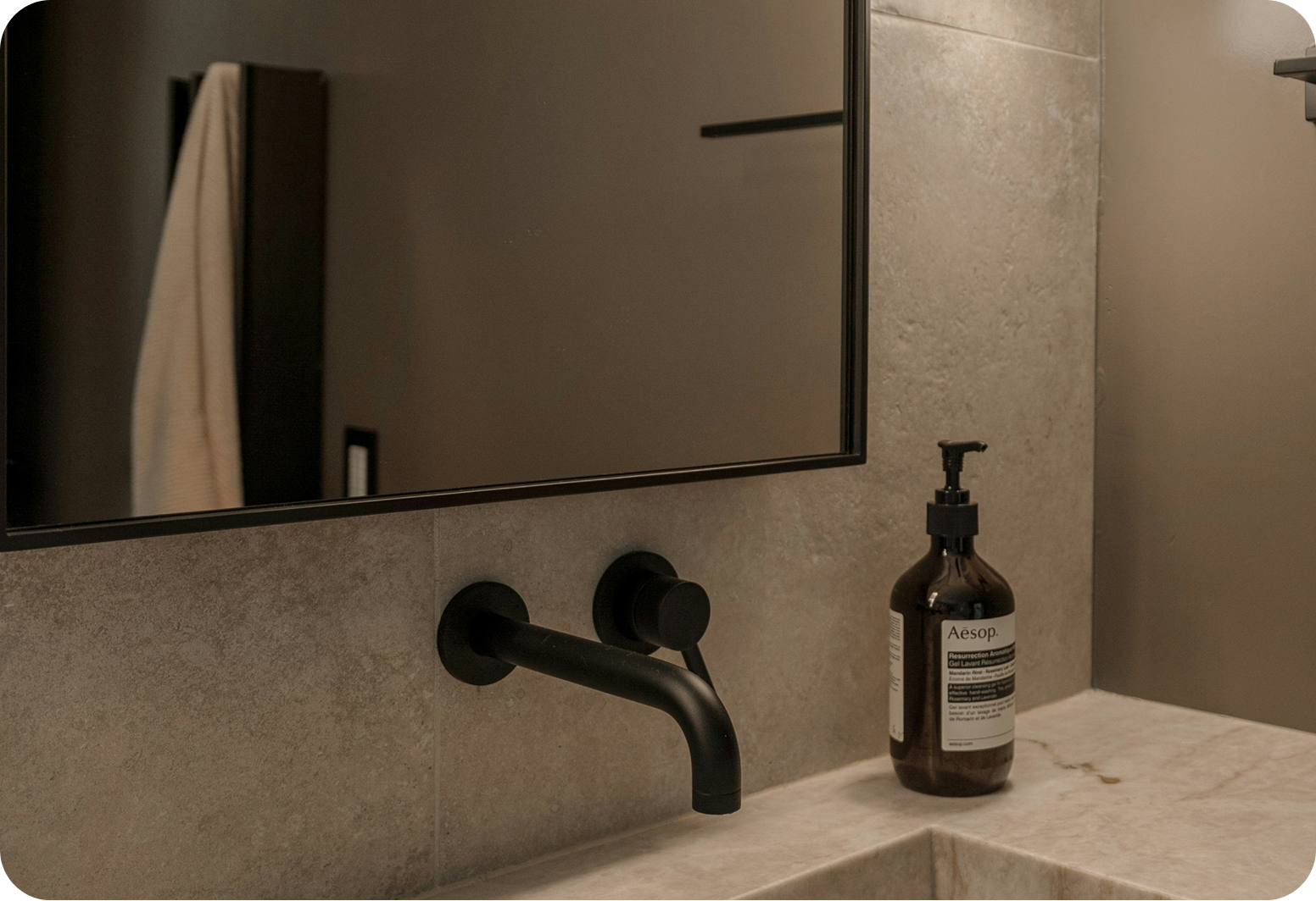 Bathroom sink with black faucet, mirror, and Aesop soap dispenser on beige marble countertop.
