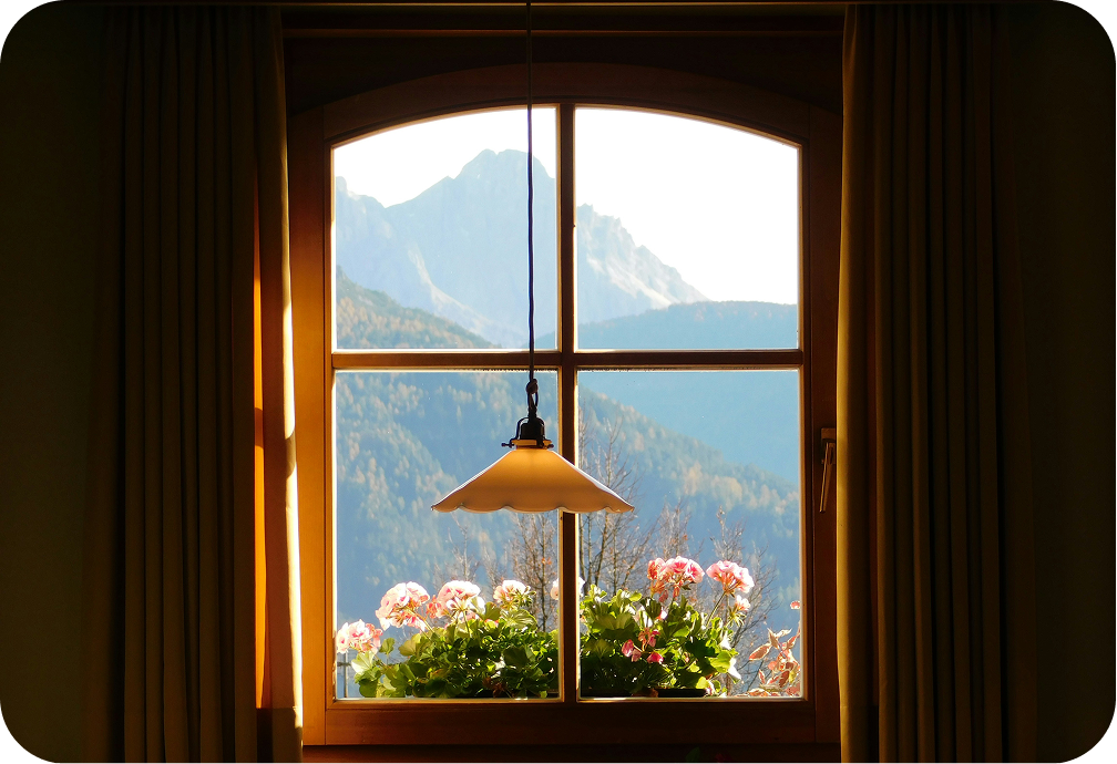 View through a window showing mountains, with potted pink and white flowers on the windowsill and curtains on either side.