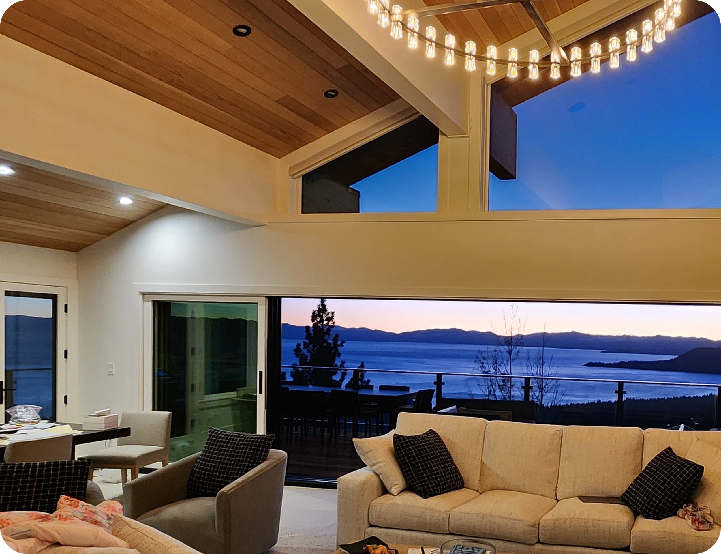 Living room with large windows showcasing a peaceful lake view at sunset. The room features a beige sofa with dark patterned pillows, a cream-colored armchair, and a dining area with black chairs. The ceiling has wooden beams and is illuminated by a string of small hanging lights. The room is decorated with modern furniture and has a cozy, elegant atmosphere.