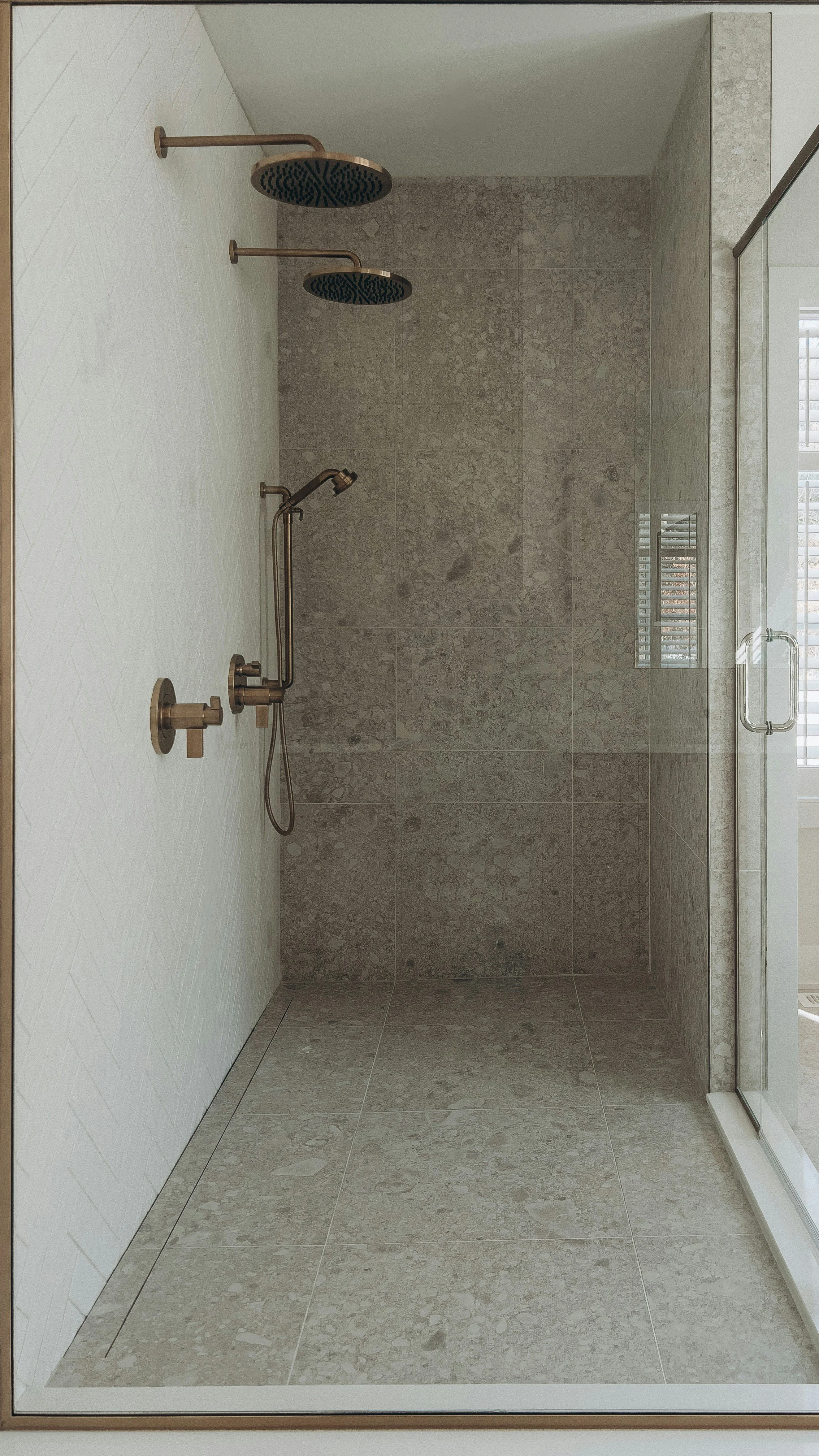 A modern walk-in shower with two rain shower heads on the ceiling, a handheld shower on the wall, beige tile flooring, and a glass door with a handle.