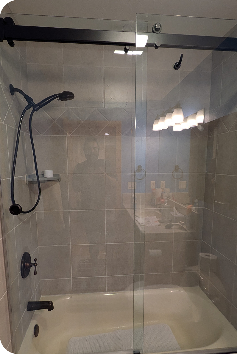 Bathroom shower with glass door, black shower fixtures, and beige tiled walls. Reflection of a person and part of a room with a mirror and light fixture visible.