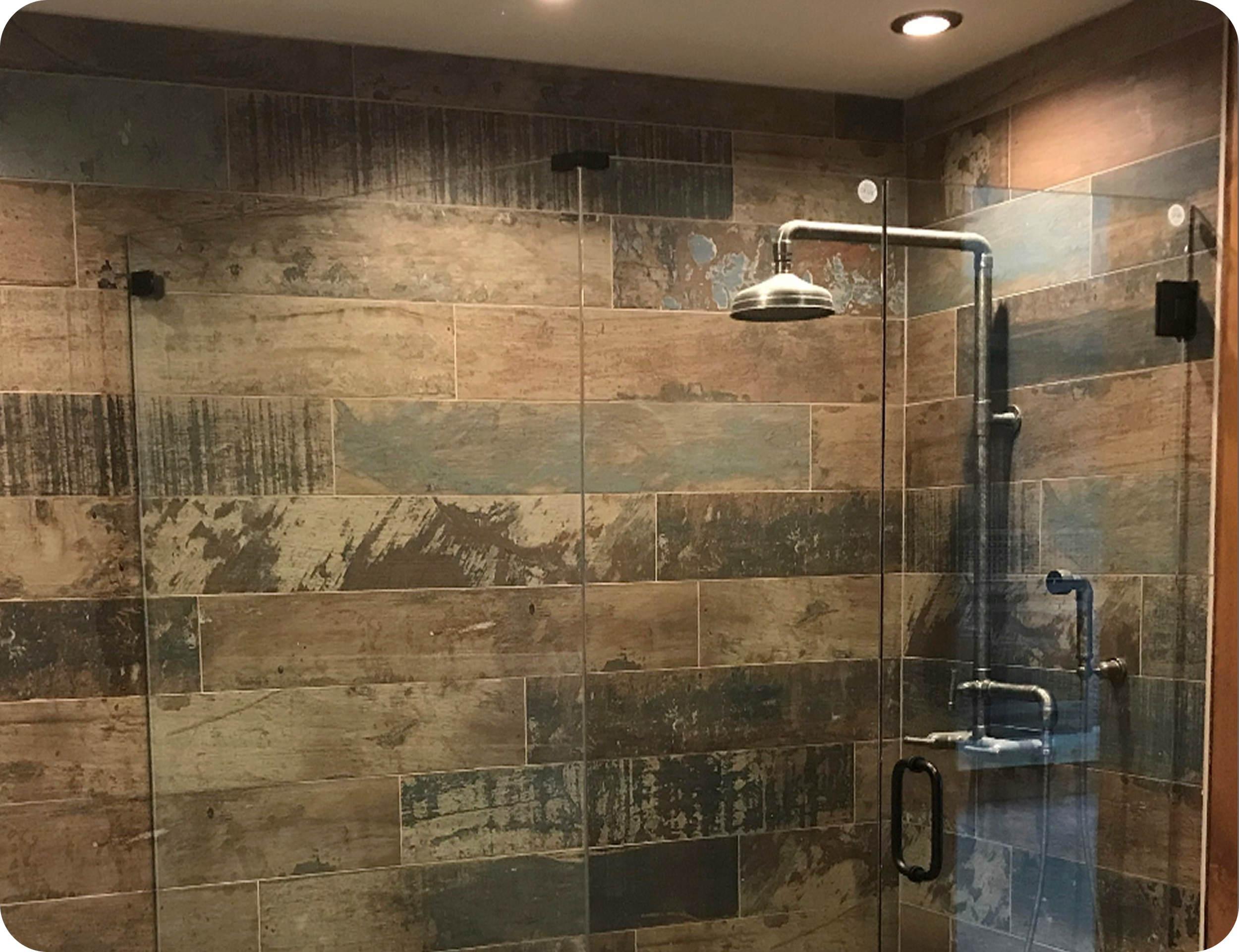 Shower with brown tile walls, overhead shower head, and glass door with black handle.