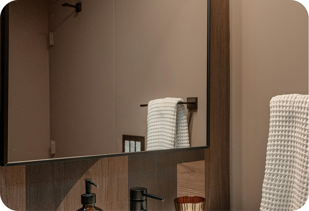 A bathroom sink area with a mirror, a dark soap dispenser, and a copper-colored cup. A white textured towel is hanging on a rack reflected in the mirror, and another similar towel is draped over a surface outside the mirror's view.