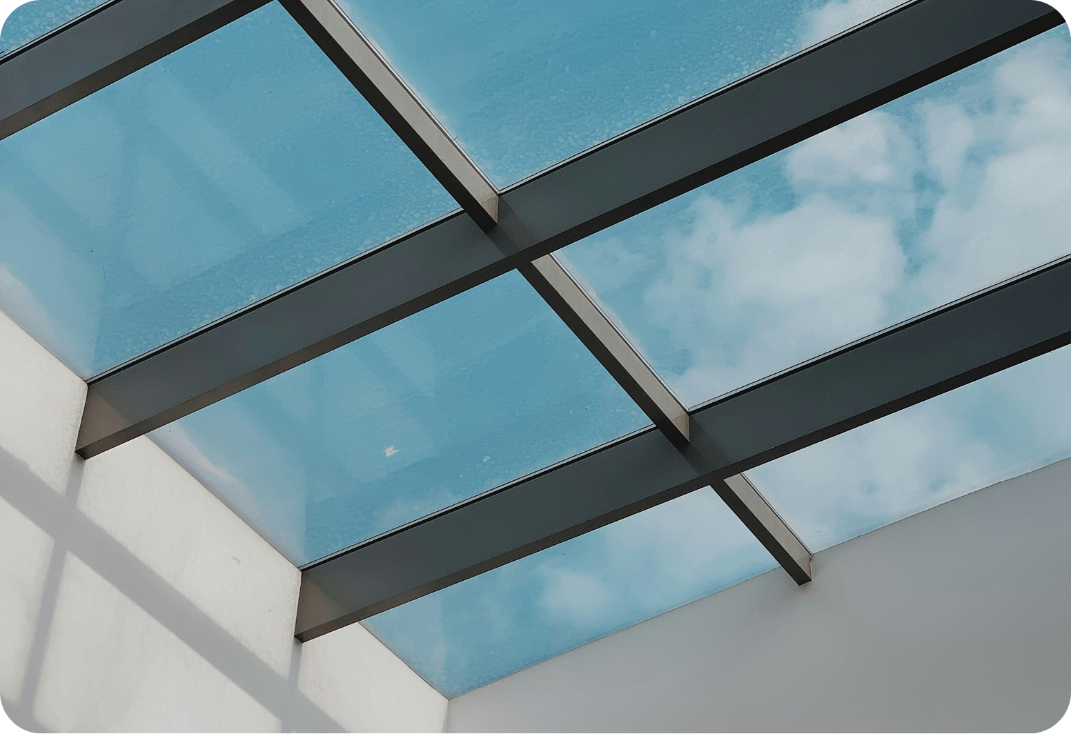 Looking up through a glass roof with metal supports at a blue sky with some clouds.