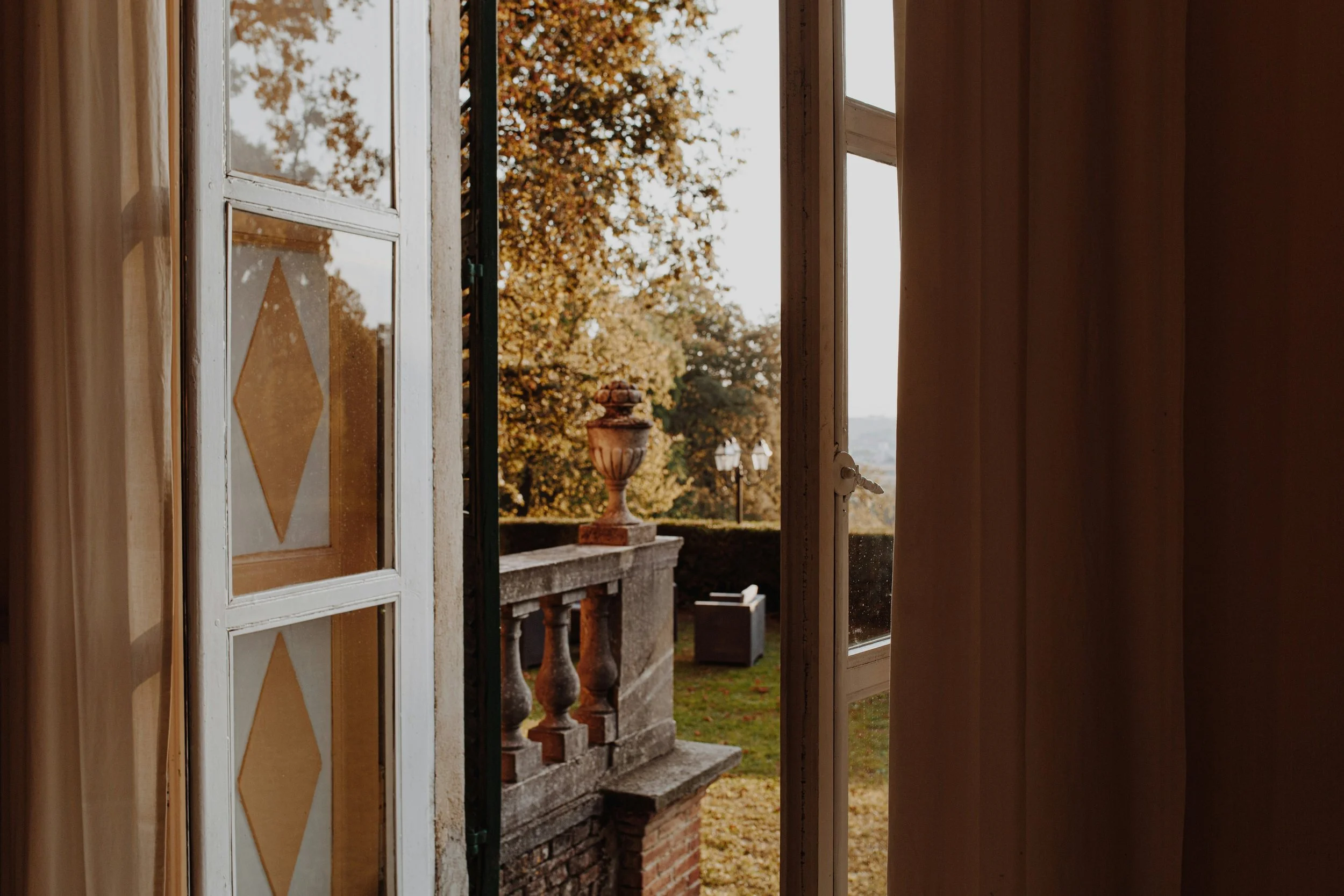 Open door leading to a balcony with a decorative stone balustrade, overlooking a garden with a large oak tree and a stone urn. The scene is set during autumn with fallen leaves.