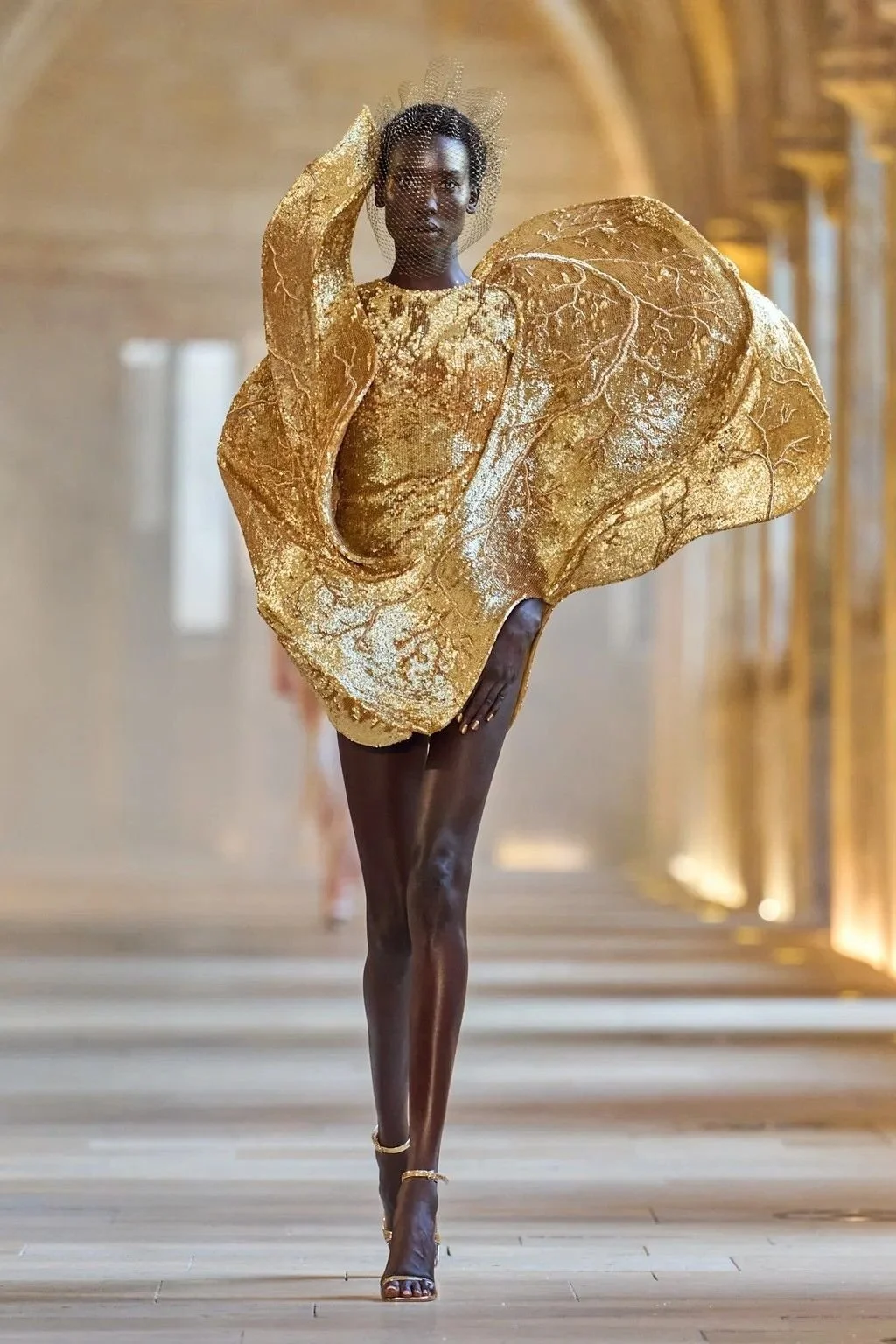 A model walking down a runway wearing a gold dress with structured, leaf-like shapes and a matching headpiece with mesh veil. luxury beauty, fragrance, skincare, haircare, fashion editorial, beauty editorial, cosmetics, cinematic, editorial, moody, h