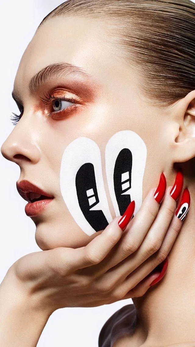 A woman with blonde hair pulled back, and makeup including bronze eyeshadow and nude lips, holding her face with her left hand. Her nails are painted red with black and white abstract designs. She has white face stickers with smiley face emoticons on her cheek.