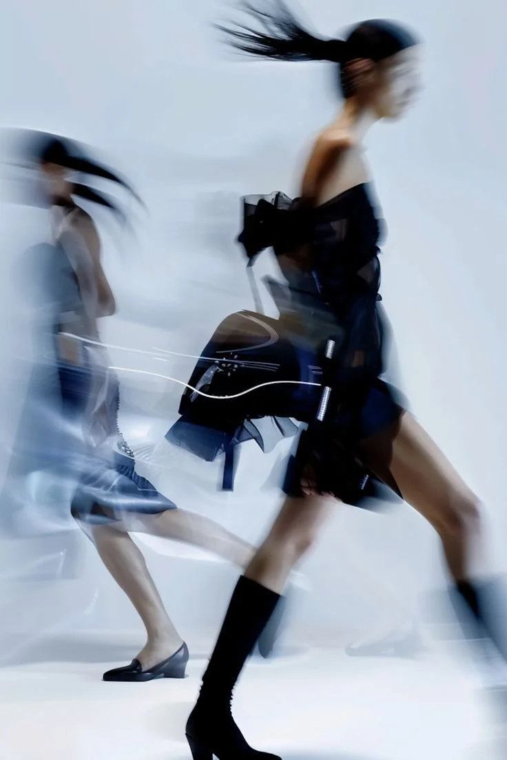 Blurry image of two women walking, one with long flowing hair, wearing a black dress, with a backpack, and black high-heeled boots, against a plain background.luxury beauty, fragrance, skincare, haircare, fashion editorial, beauty editorial, cosmetic
