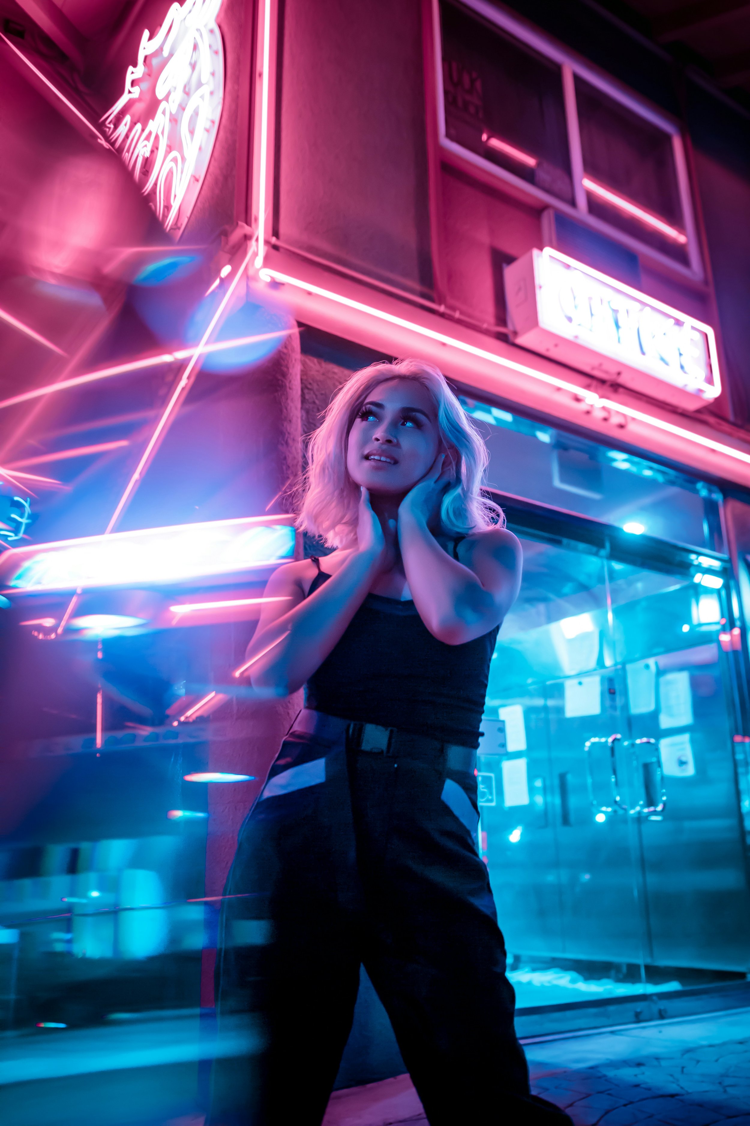 A woman with blonde hair standing in front of neon signs that say 'Bar', 'Wine', and 'Liquor' with colorful pink, blue, and purple lighting reflections.