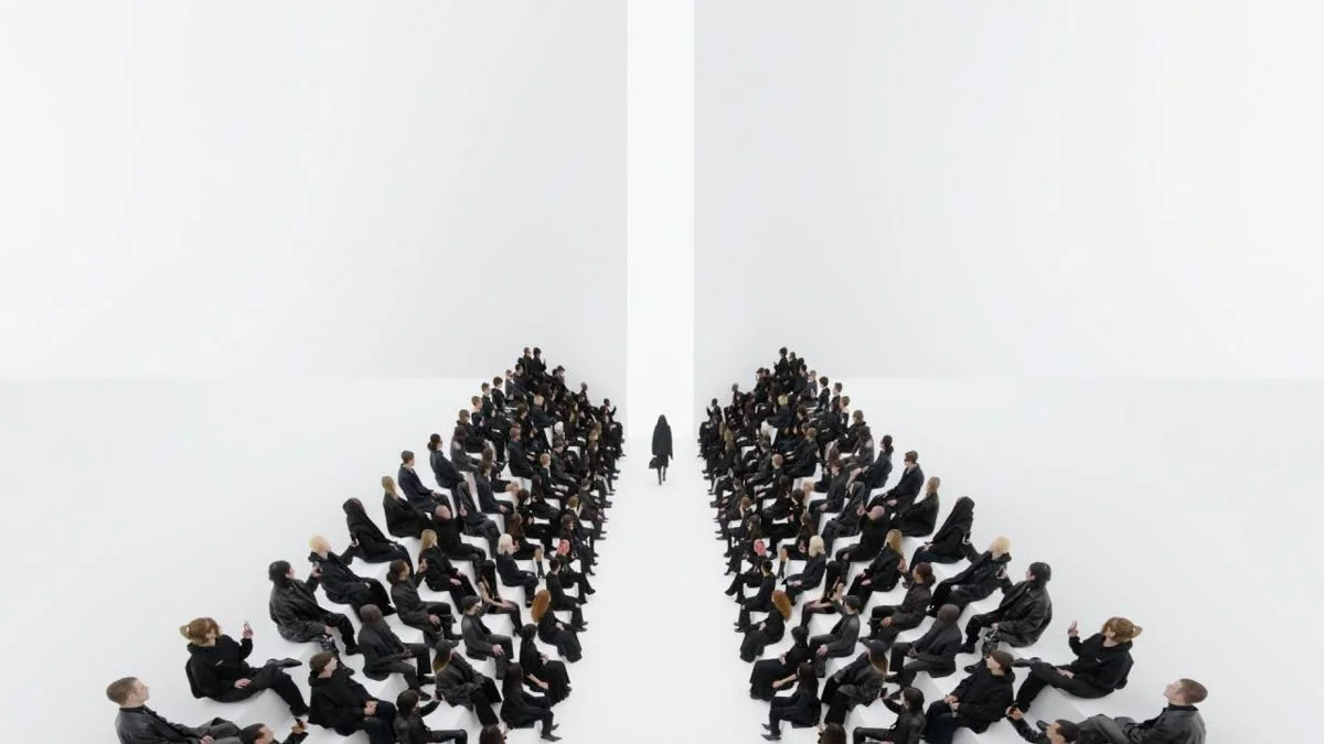 A large group of people dressed in black suits are seated in two symmetrical rows facing each other in a white room, with one person walking toward the back of the room. luxury beauty, fragrance, skincare, haircare, fashion editorial, beauty editoria