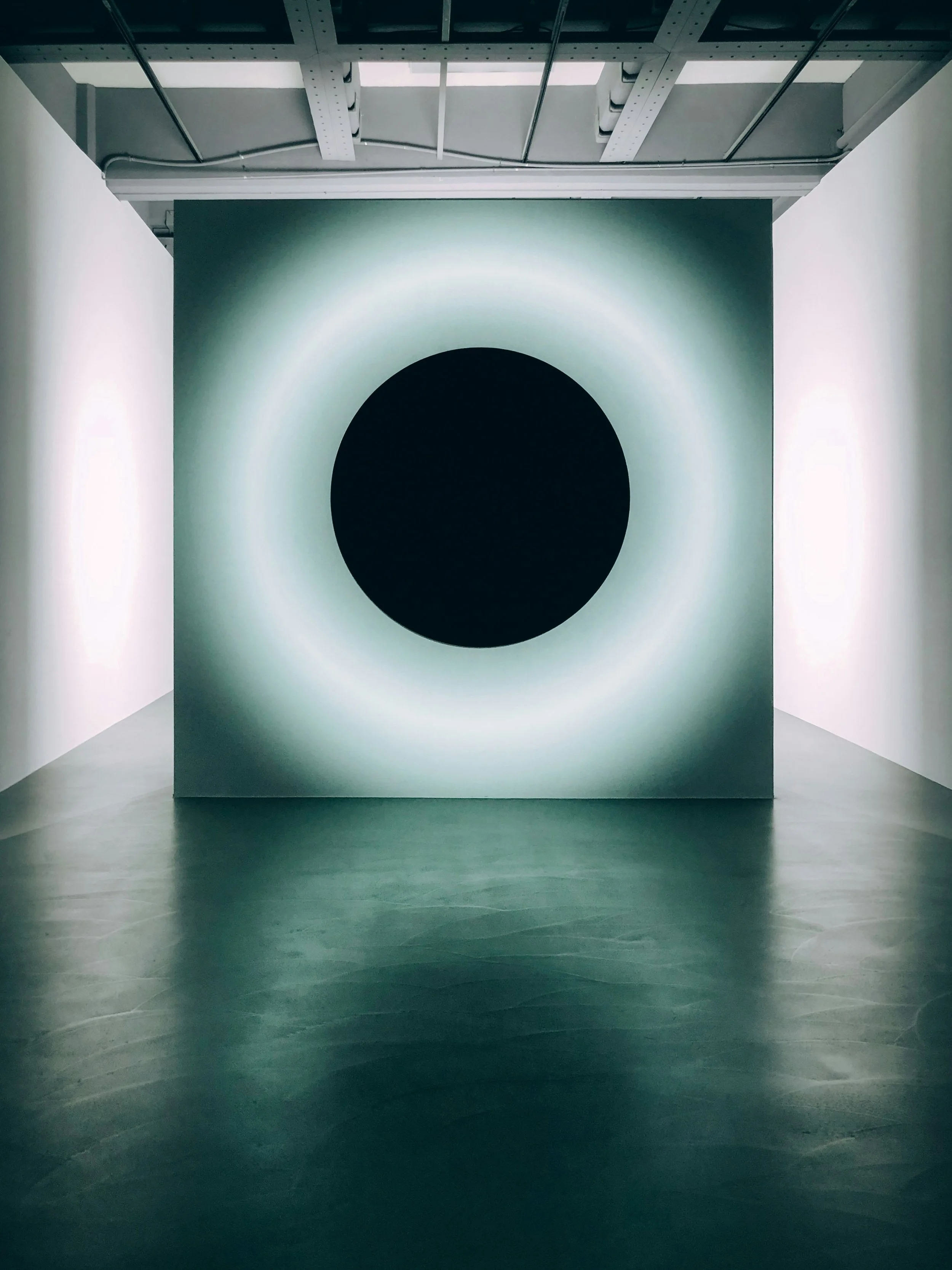 An art installation with a circular black hole at the center, surrounded by a glowing ring of light, in a white-walled room with a gray ceiling and wooden floor.