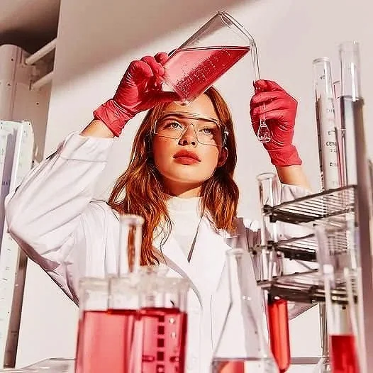 A female scientist in a white lab coat and red gloves pouring a pink liquid from a beaker into a test tube in a laboratory setting.