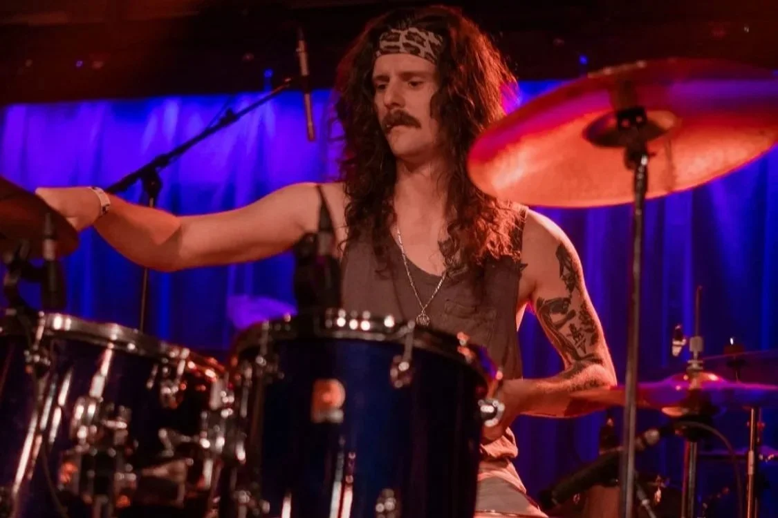 A drummer with long curly hair, a mustache, and tattoos is playing drums on a stage with purple curtains behind him.