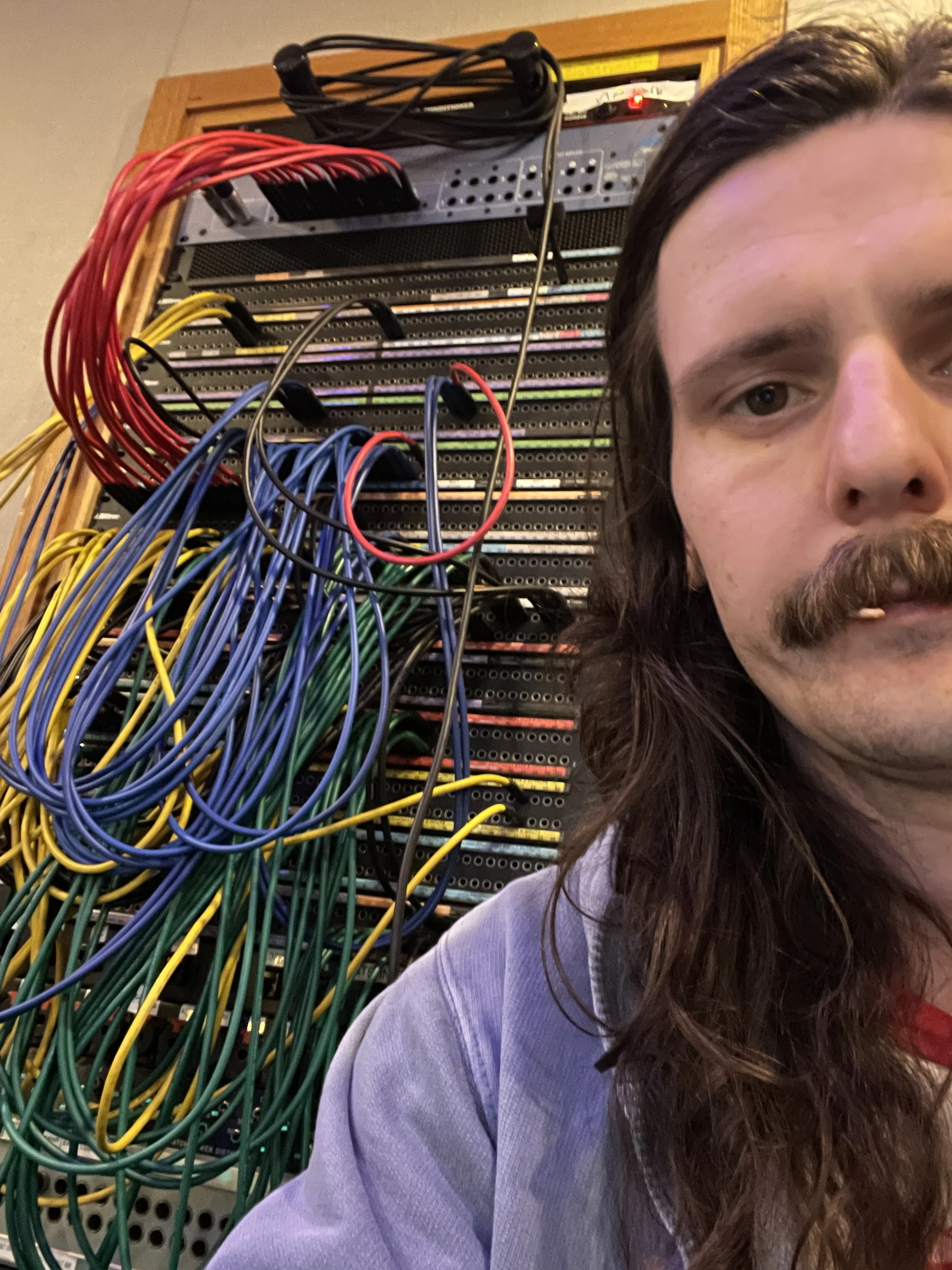 A person with long brown hair and a mustache sitting in front of a server rack filled with colorful network cables.