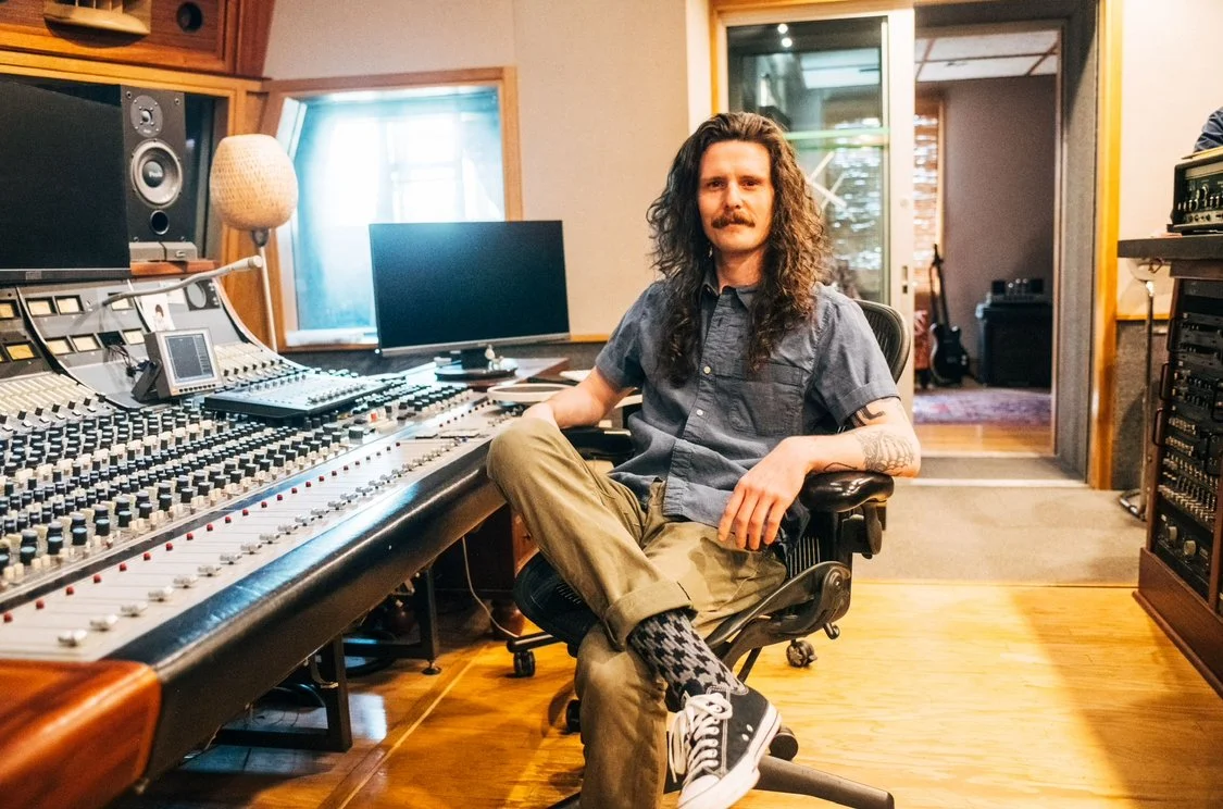 A man with long hair and a mustache sitting in a recording studio.