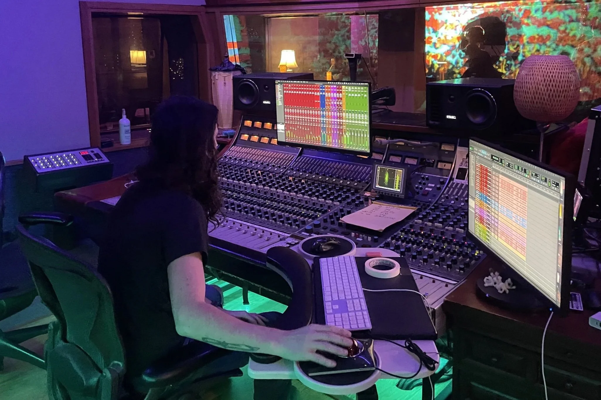 A music recording studio with a person working at a large mixing console, surrounded by multiple computer screens displaying mixing software, large studio speakers, and a window showing another room with colorful projections and people inside.