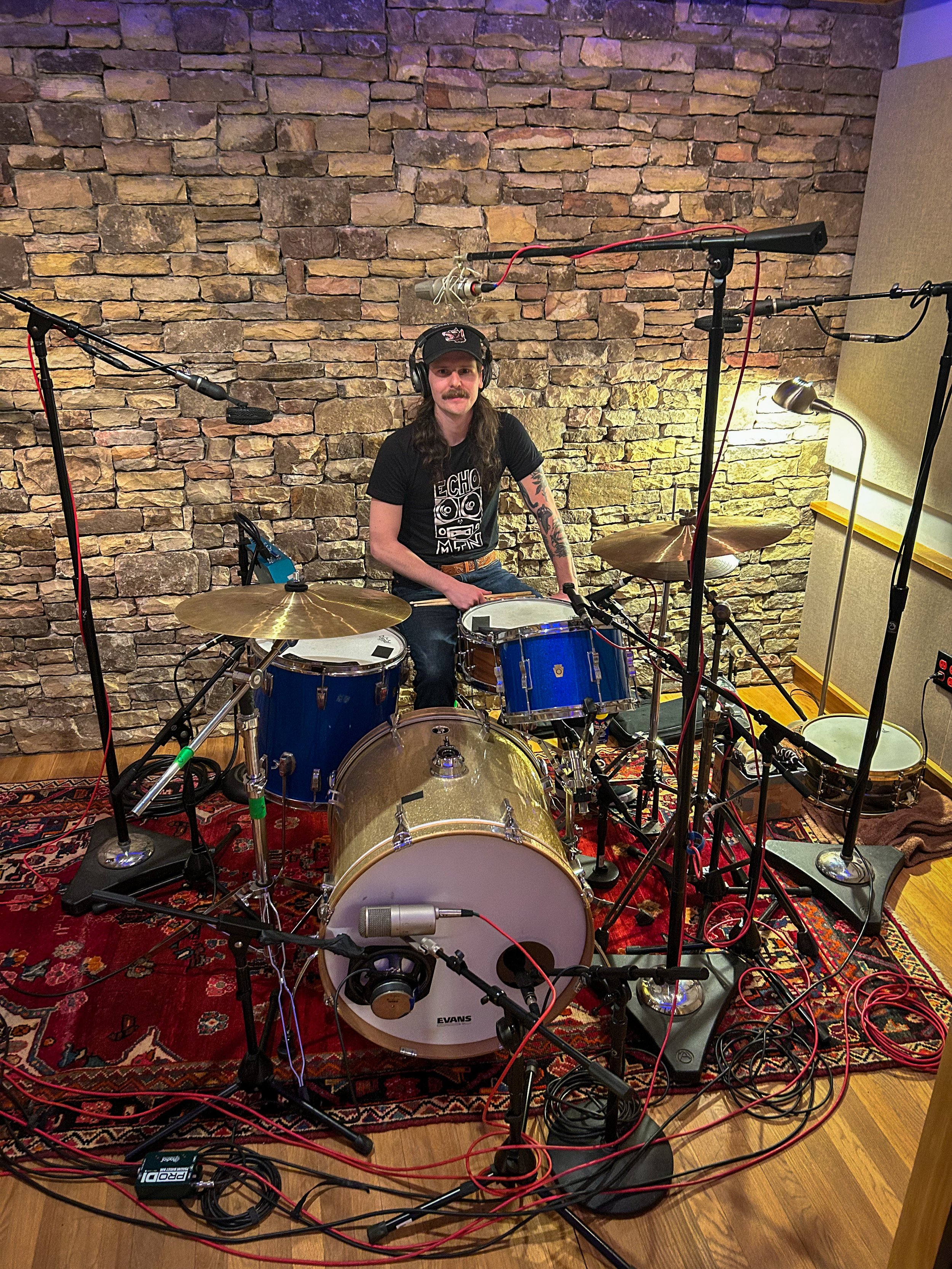 A man with long hair and a mustache sitting at a drum set in a recording studio with a stone wall, surrounded by microphones and cables, wearing a black T-shirt with a graphic and a black cap.
