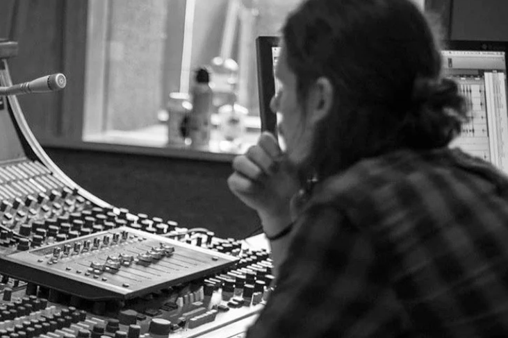 A person with dark curly hair tied back, sitting at a sound mixing board in a recording studio, looking at computer monitors.