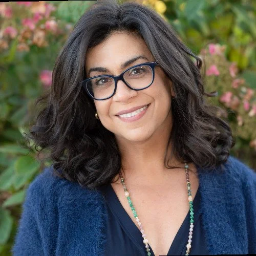 Close-up of a woman with dark hair, glasses, and a bright smile, wearing a blue top and a gold necklace.