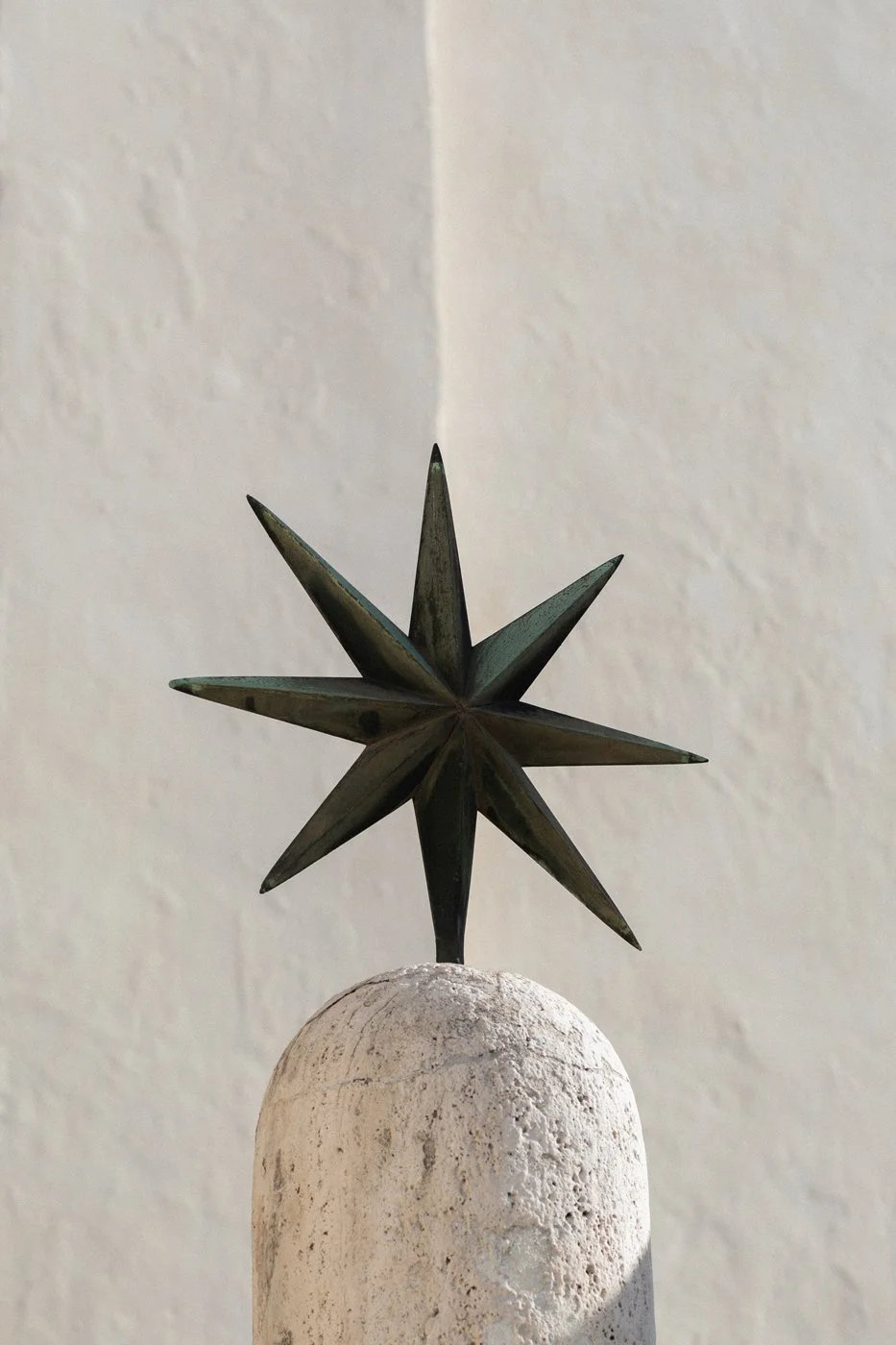 A decorative metal star sculpture with a rustic finish on a white, textured stone pillar against a plain wall.