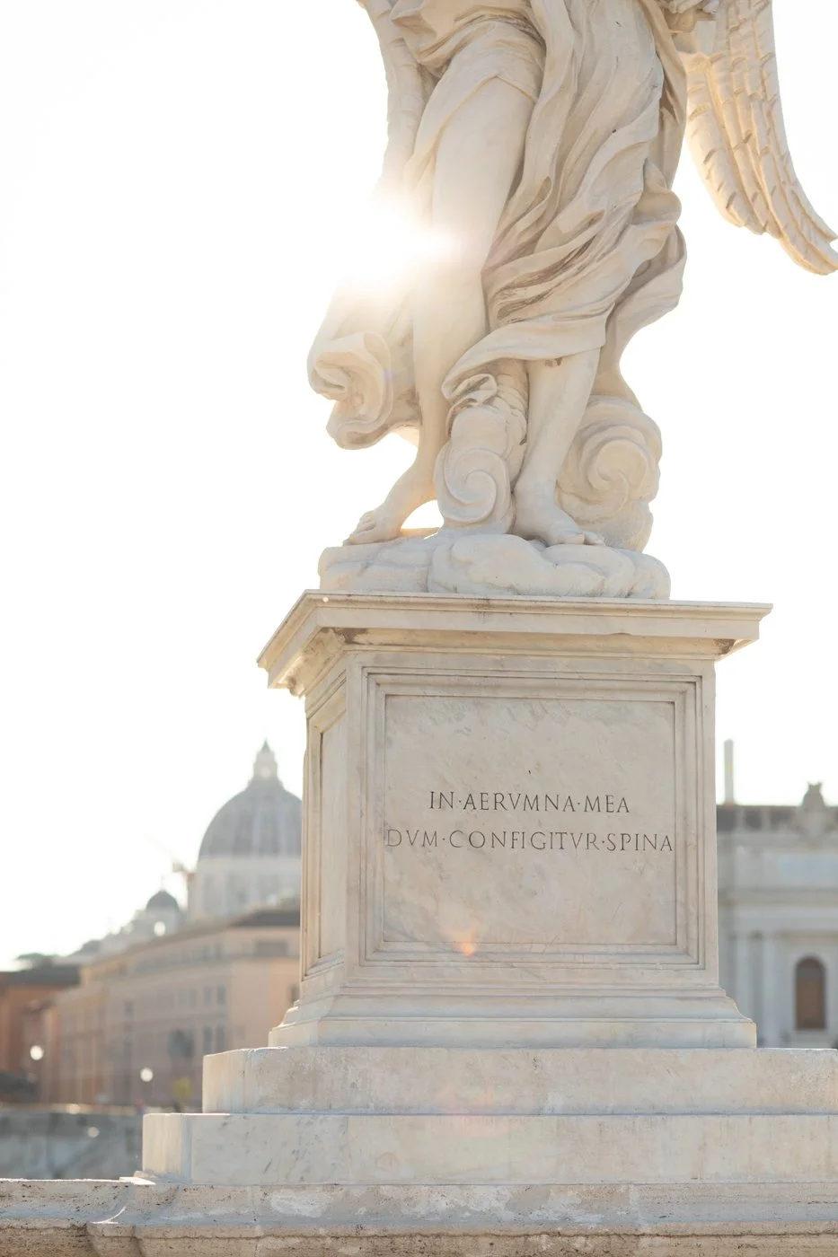 Close-up of a white marble statue on a pedestal with an inscription, with a cityscape and a domed building in the background, illuminated by sunlight.