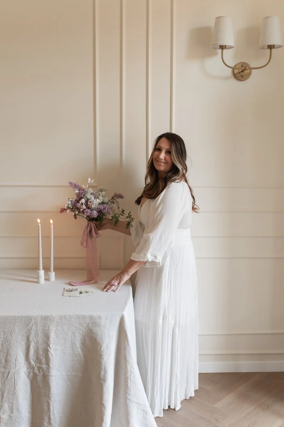 A woman in a white dress stands next to a table with lit candles and a bouquet of pink and purple flowers, in a softly lit room with beige walls and a wall sconce.