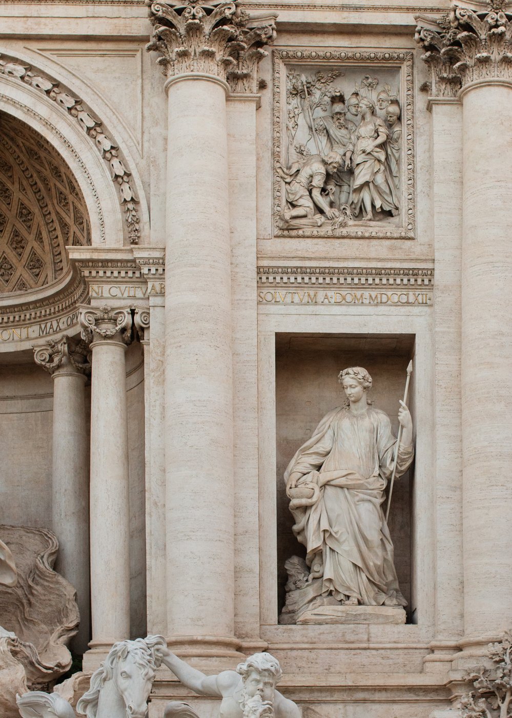 Sculpture of a seated woman holding a staff, with draped clothing, in an architectural niche; additional carvings and columns are visible in the background.