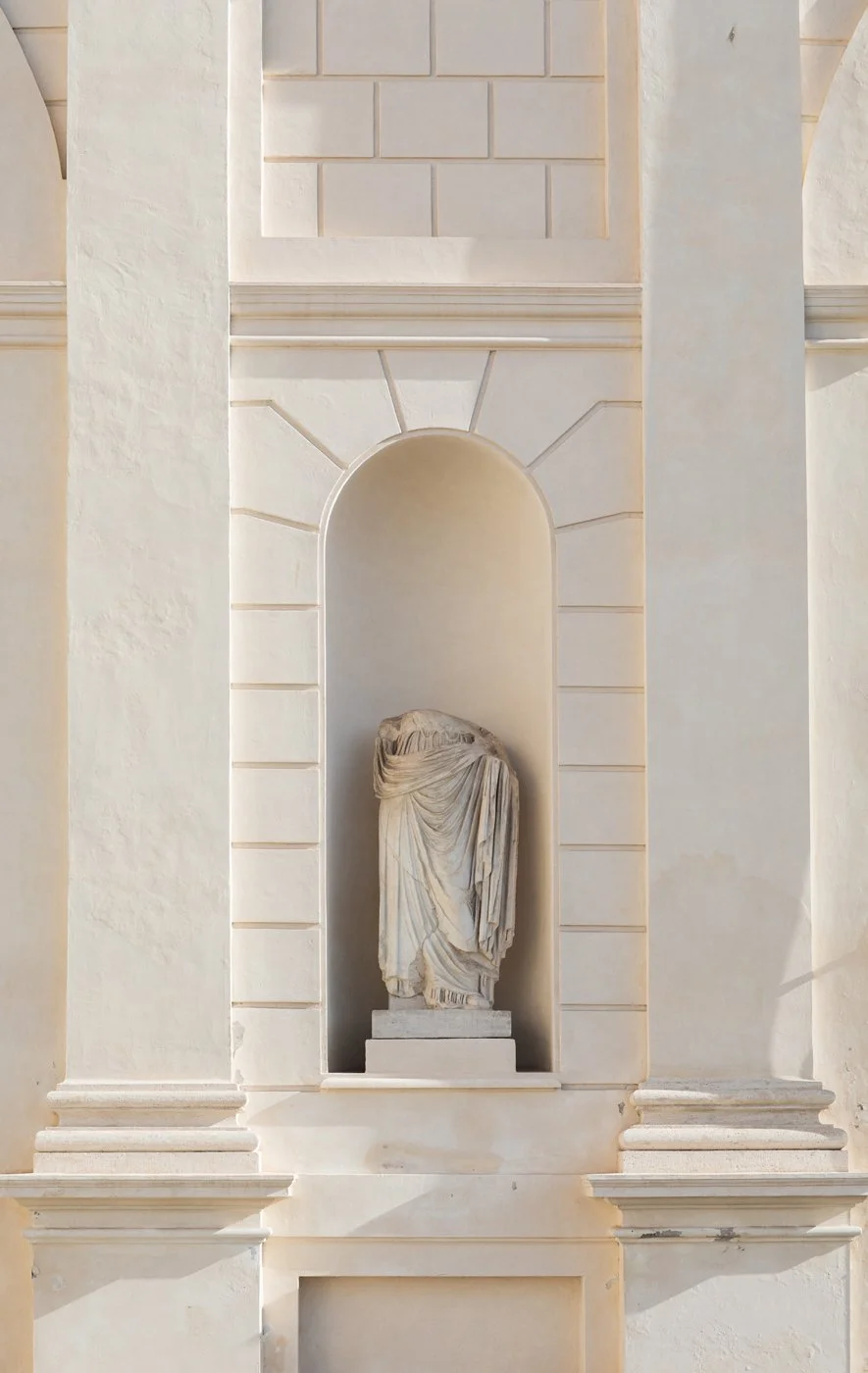 A white marble statue of a draped figure nestled in a niche in a white wall with architectural details.