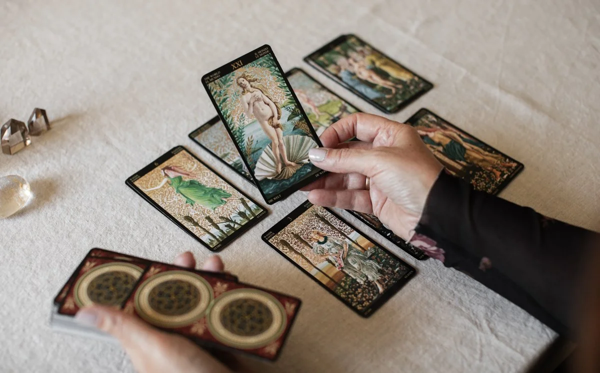 A person's hands holding tarot cards over a beige fabric surface, surrounded by more cards and small decorative objects.