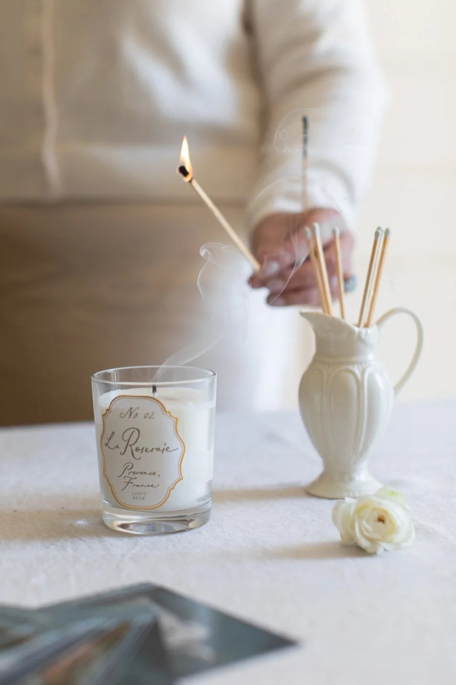 A person is lighting a matchstick inside a white candle on a table. The table has a white tablecloth, a white ceramic pitcher with several unused matchsticks, and white roses. The candle has a label that reads "La Roseraie, Provence, France." The background is softly lit.