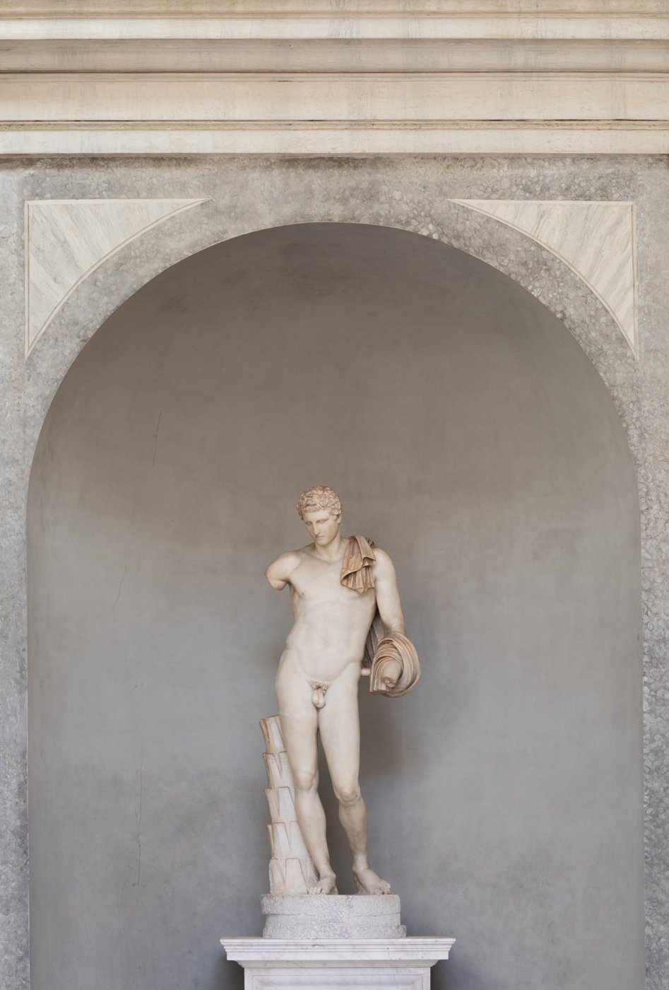 Marble sculpture of a young male nude figure with a draped cloth over his shoulder, standing on a pedestal against a gray wall within an arch.