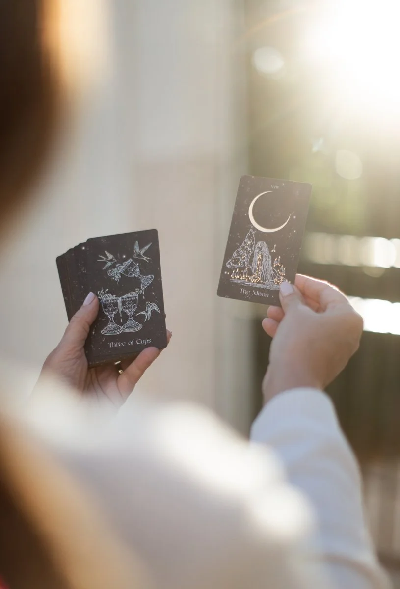 Two tarot cards being held by a person, with one card showing a moon and two wolves and titled 'The Moon,' and the other card showing two cups being poured over each other and titled 'Three of Cups.'