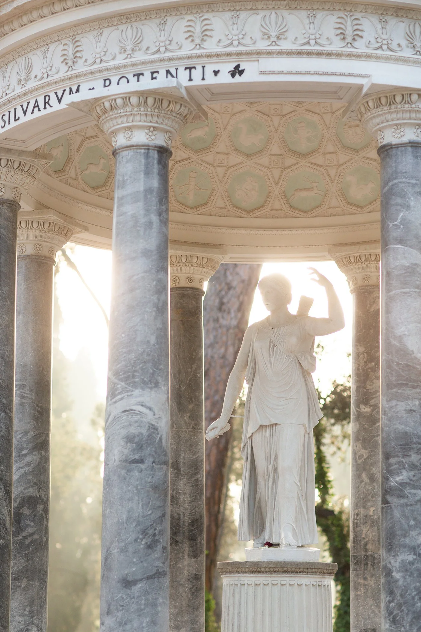 A classical marble statue of a woman standing inside an ornate gazebo with Corinthian columns and decorative friezes, sunlight shining through trees in the background.
