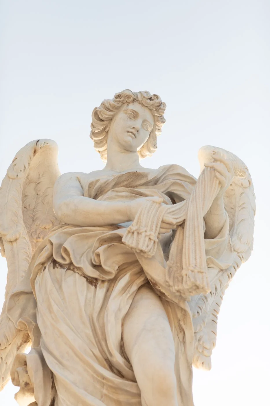Close-up of a marble angel statue with detailed wings, holding a cloth, against a clear sky.