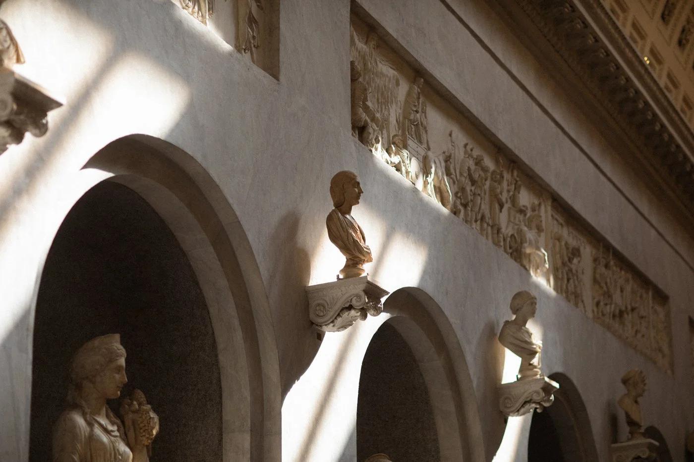 Photograph of classical sculptures of women, lined up in arched niches on a wall, with sunlight casting shadows on them.