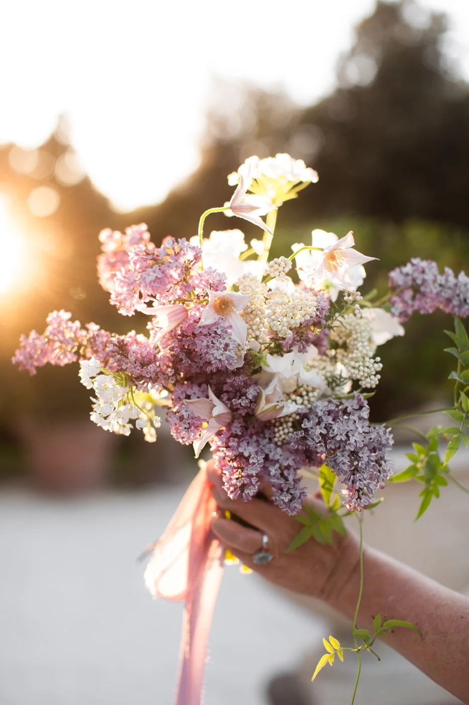 A hand holding a bouquet of pink, white, and purple flowers with a sunset in the background.