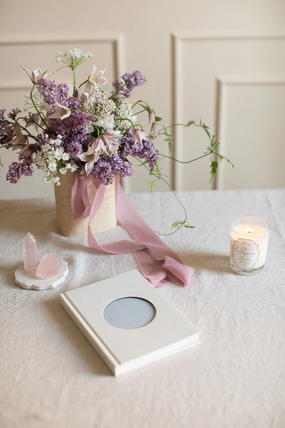 A floral arrangement of lavender and white flowers in a beige vase, tied with a pink ribbon, on a white table. Nearby are a lit candle in a glass holder with a label, a small white dish with pink crystals, and a closed white notebook with a circular mirror on the cover, set against a light-colored wall with paneled decor.