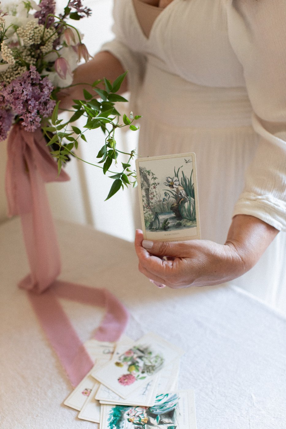A woman in a white dress holding a vintage style card with plants and animals, with floral cards on a table and a flower arrangement with greenery and pink flowers nearby.