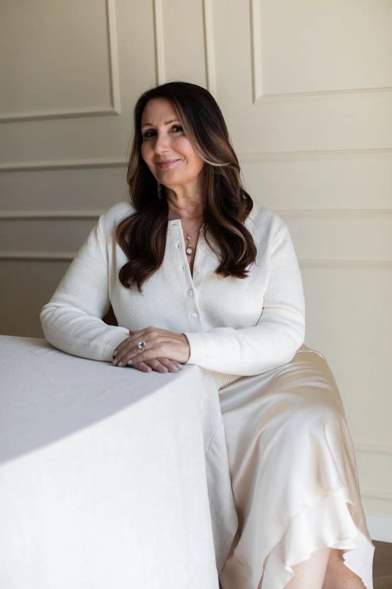 A woman with long, dark brown hair with lighter highlights, wearing a white cardigan, sitting at a table with a cream-colored tablecloth, smiling softly at the camera in front of cream paneled walls.