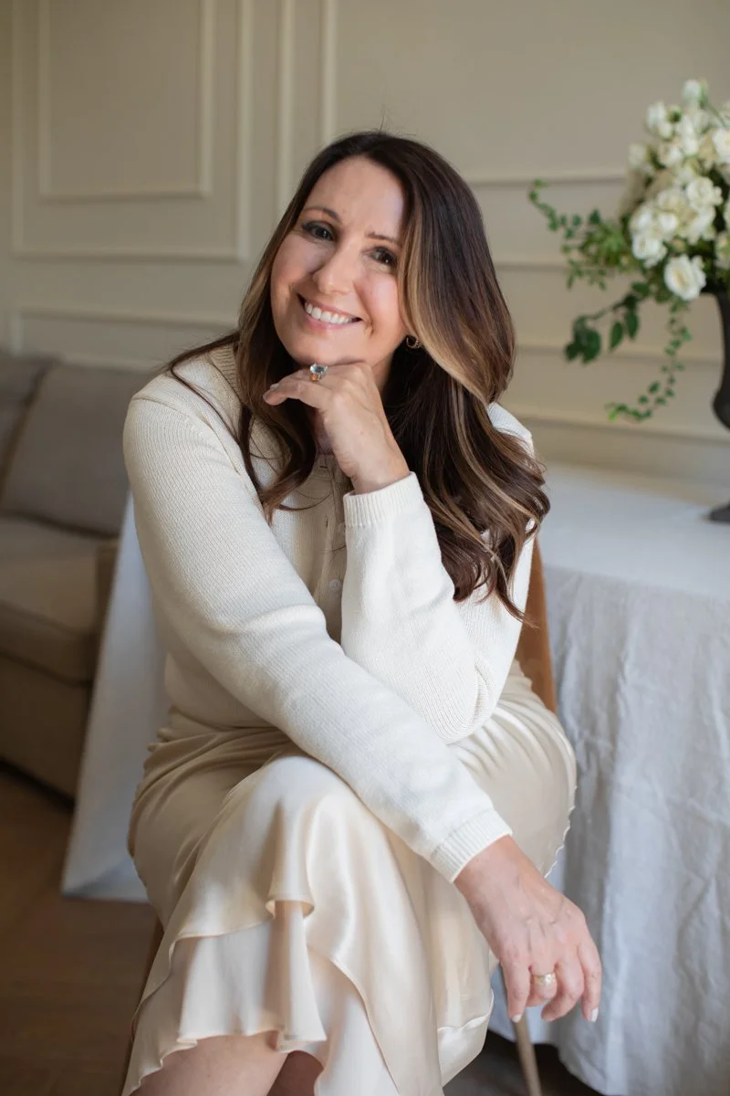 A woman with long brown hair smiling and resting her chin on her hand, sitting in a cozy room with cream-colored walls, a beige sofa, and a table with a white cloth and a vase of white flowers.