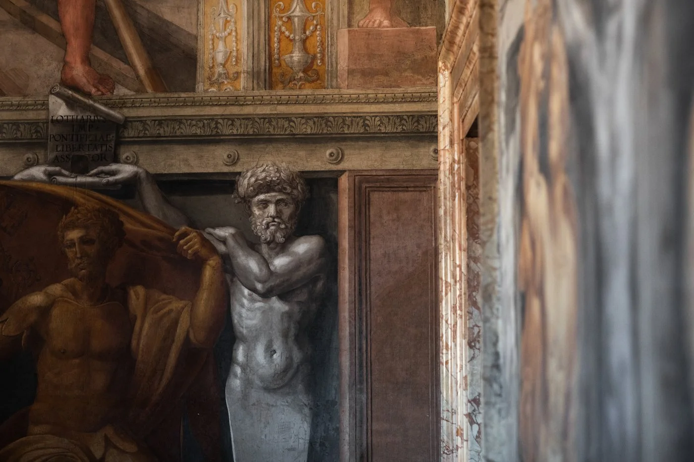 Close-up of a classical religious or historical sculpture depicting a bearded man with curly hair and muscular torso, holding a mantle or cloth. Part of a larger scene with other figures and intricate architectural and decorative elements in the back