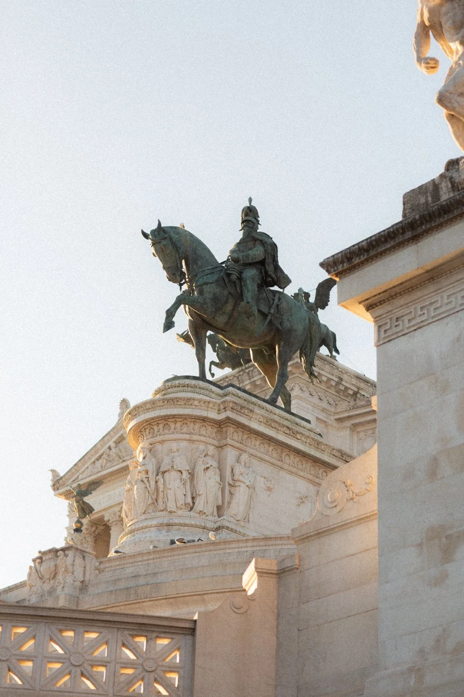 Close-up of a bronze equestrian statue atop a white stone pedestal, featuring detailed carvings and figures, with a clear sky in the background.
