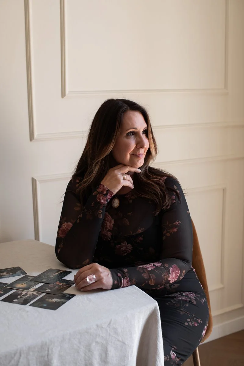 A woman with long brown hair, wearing a dark floral dress, sitting at a table with photographs spread out in front of her, posing with her chin resting on her hand and looking to the right.